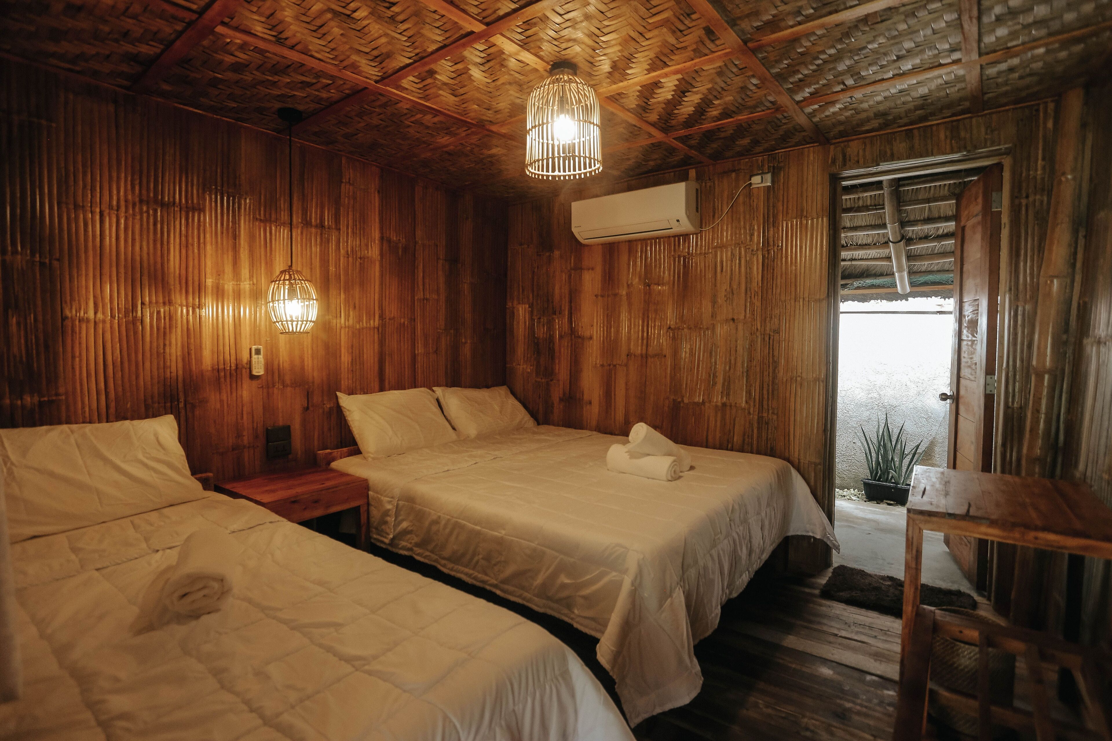 Una habitación con dos camas, paredes de madera, techo tejido y una lámpara colgante.