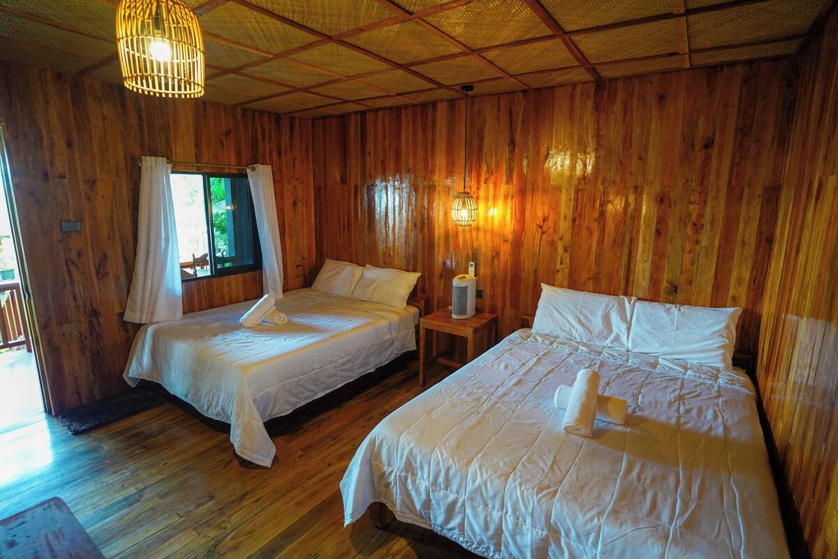 Una habitación de cabaña de madera con dos camas, una ventana con cortinas, una lámpara colgante y una mesita con lámpara.