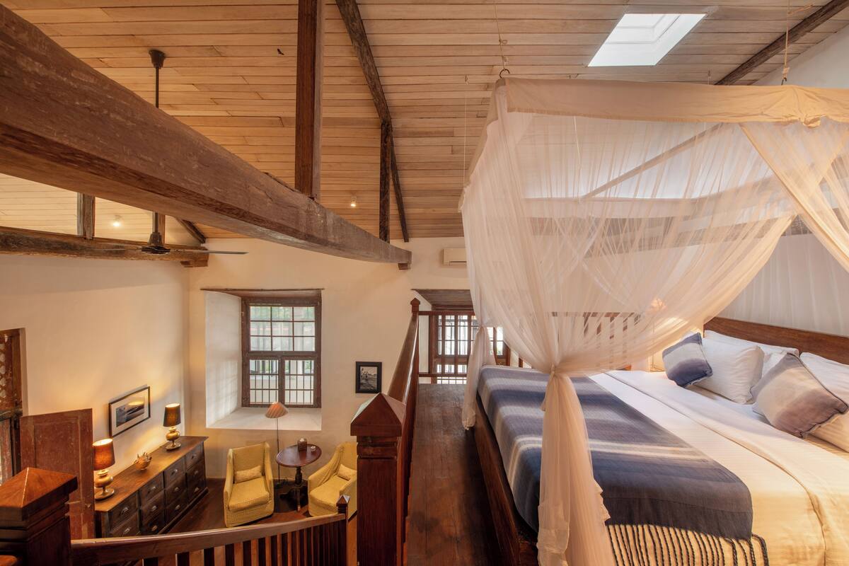 A bedroom with a canopy bed, wooden beams, a desk with a chair, and a window with barred glass.