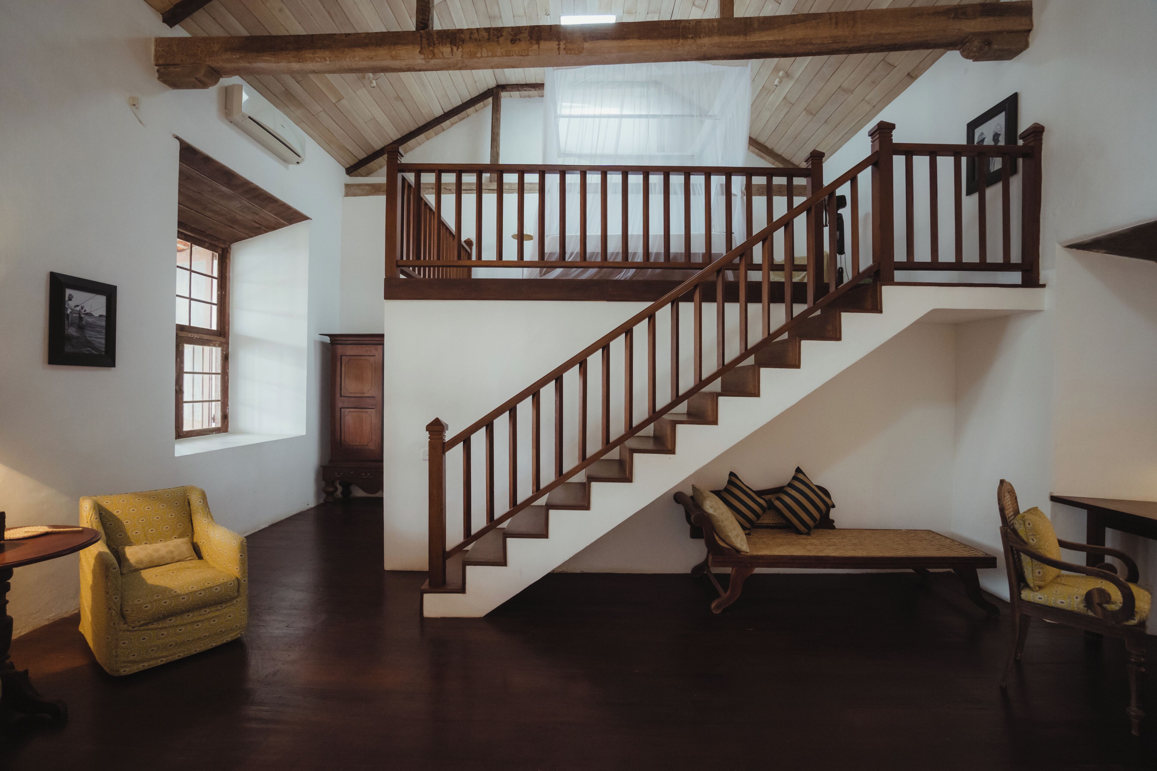 Eine Treppe mit hölzernen Geländern führt zu einer oberen Etage, ein Holzstuhl, eine Bank und ein gerahmtes Bild an der Wand.
