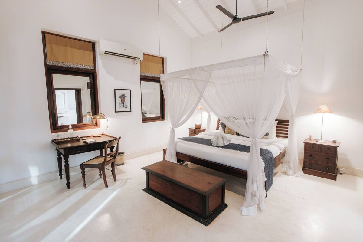 A bedroom with a four-poster bed, a desk, a chair, a bedside table, and a nightstand.