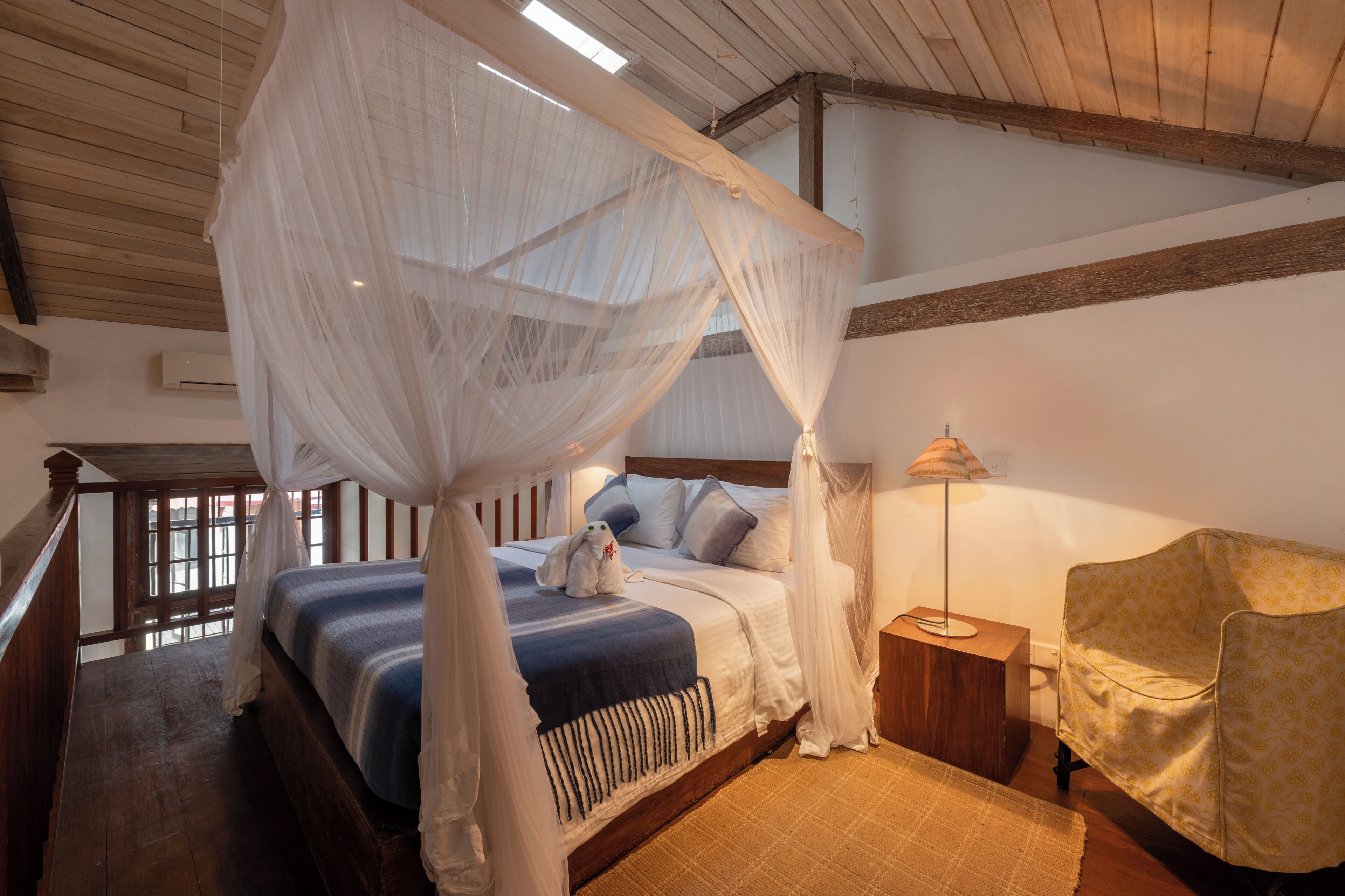 Un dormitorio con cama de dos columnas y dosel, suelo de madera, mesita de noche con lámpara y sillón estampado.