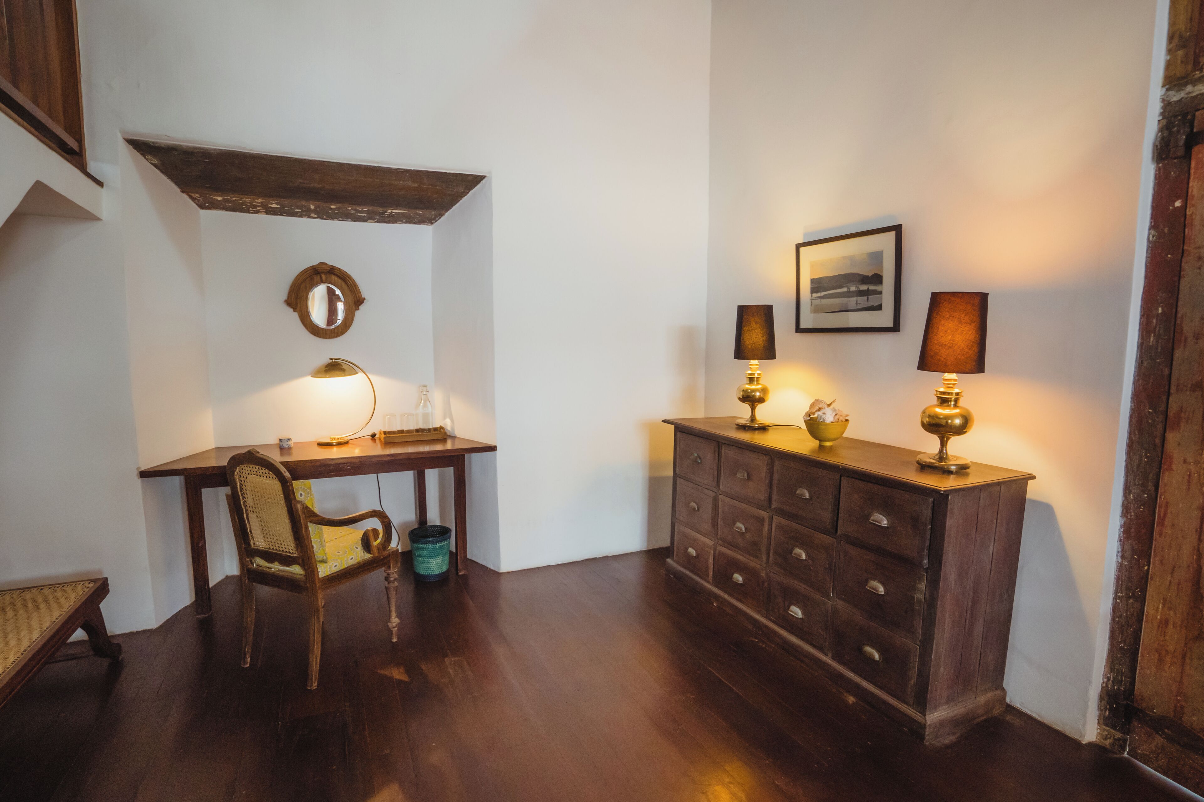 A room with a wooden desk, a chair, a dresser with drawers, a mirror, and two wall-mounted lamps.