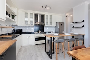 A modern kitchen with white cabinets, a stainless steel refrigerator, a wooden table, and bar stools. - Searenity (Cape Town)