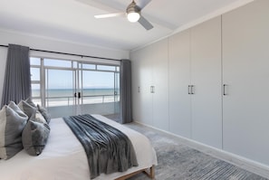 A bedroom with a large bed, a ceiling fan, and a view of the ocean through a large window. - Searenity (Cape Town)