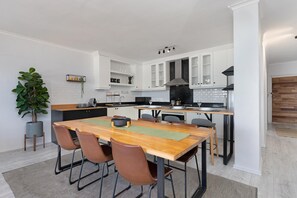 A modern kitchen with white cabinets, a wooden dining table, and a large plant. - Searenity (Cape Town)