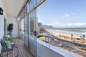 A balcony with a view of the beach and ocean, featuring a seating area with chairs and a table, and a potted plant. - Searenity (Cape Town)