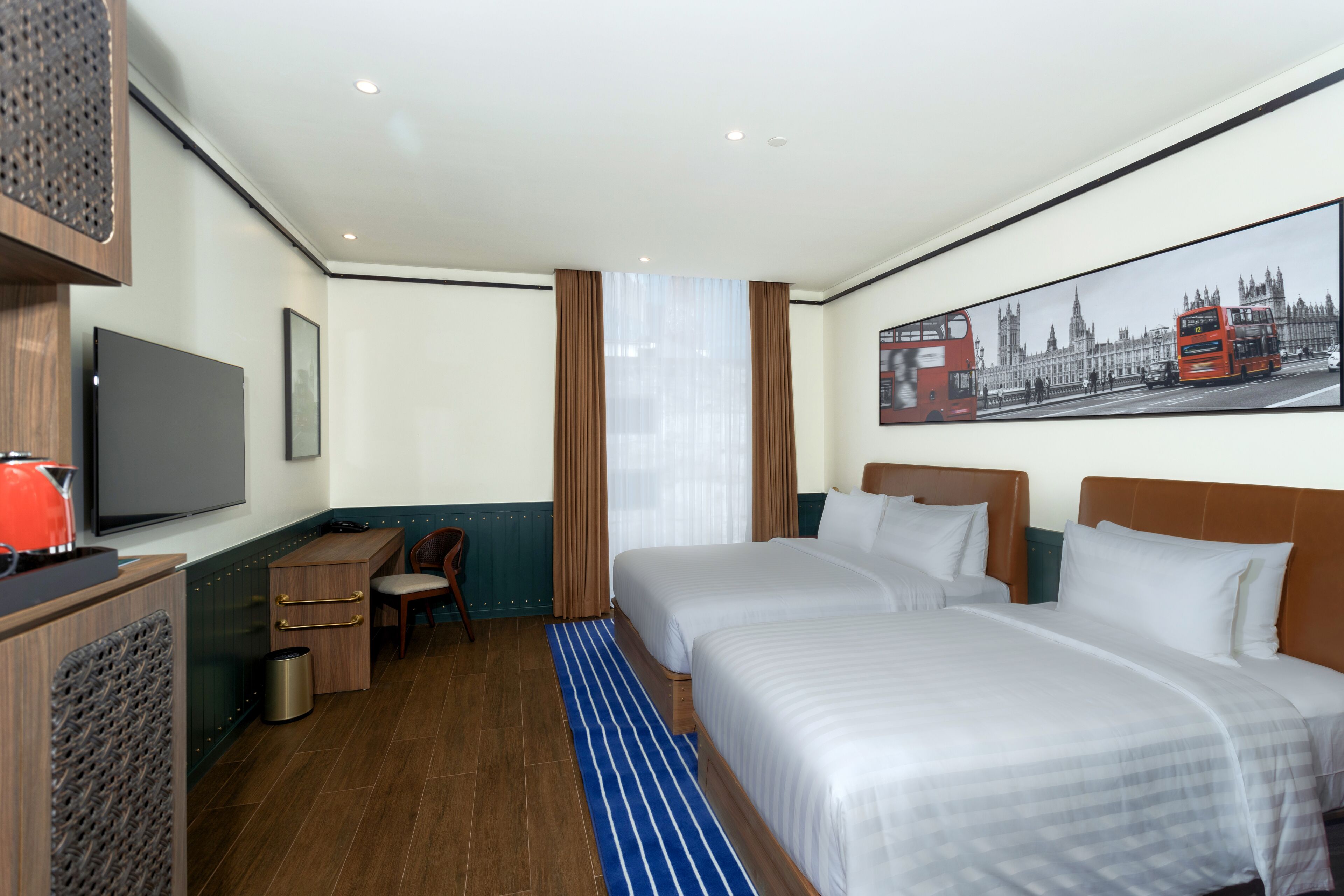 Habitación de hotel con dos camas, televisión, escritorio y un cuadro de un autobús rojo de dos pisos.