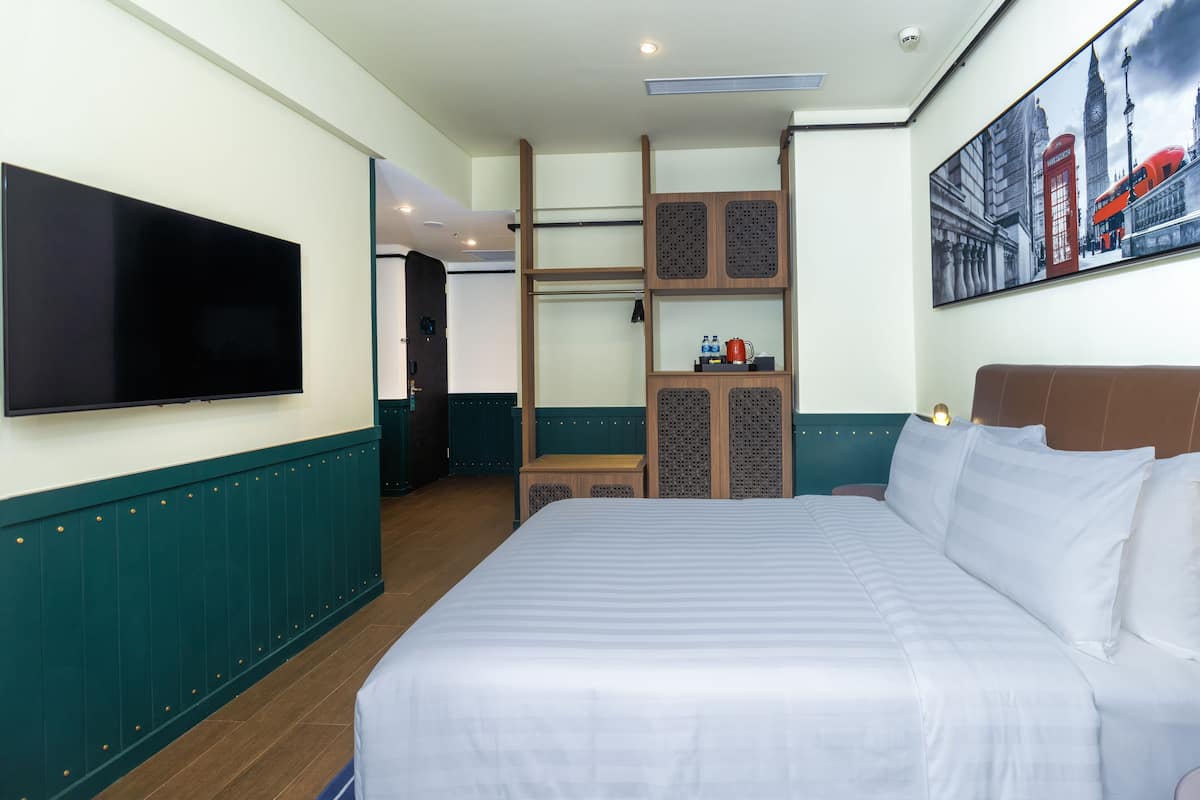 Une chambre d’hôtel avec un grand lit, un téléviseur à écran plat fixé au mur, un tableau représentant un paysage urbain et une étagère en bois avec une bouilloire et des bouteilles d’eau.