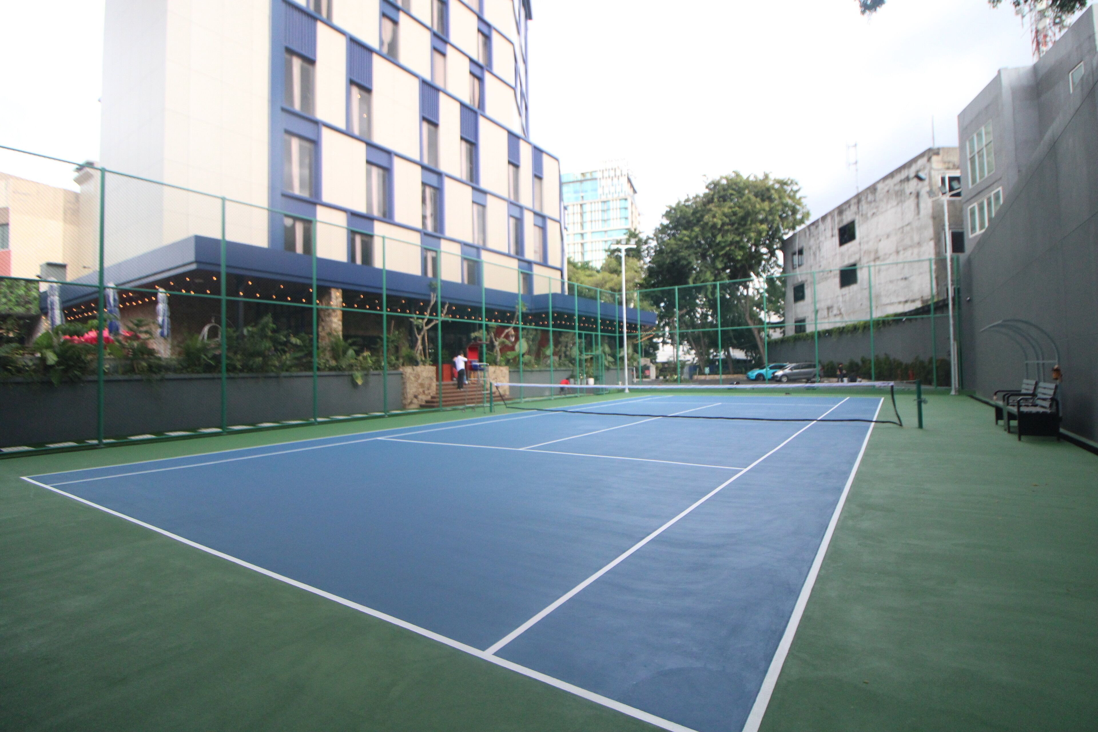 An outdoor tennis court with a net, surrounded by a green fence and a building in the background.