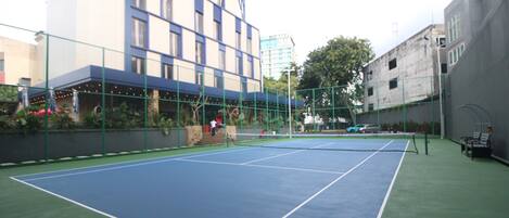 An outdoor tennis court with a net, surrounded by a green fence and a building in the background.