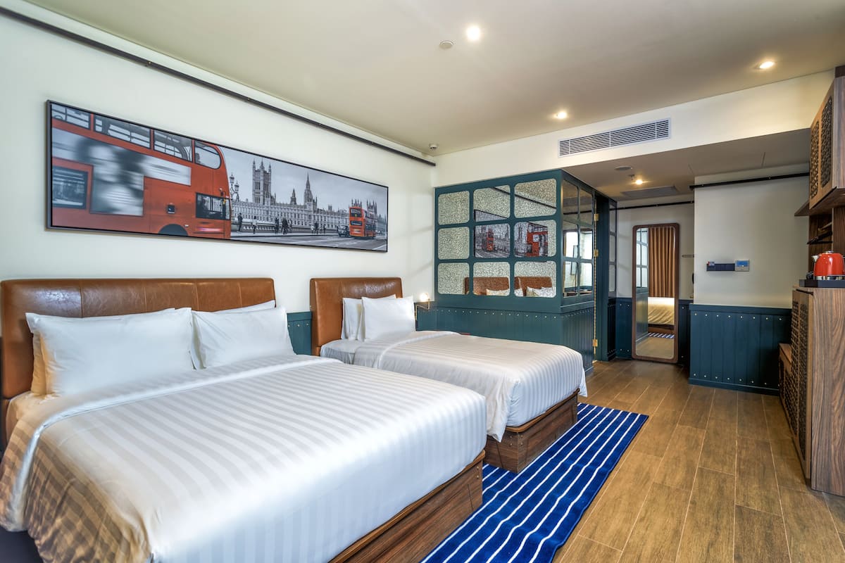 Une chambre d’hôtel avec deux lits, un grand tableau représentant un paysage urbain, une tête de lit en bois et un tapis à rayures bleues.