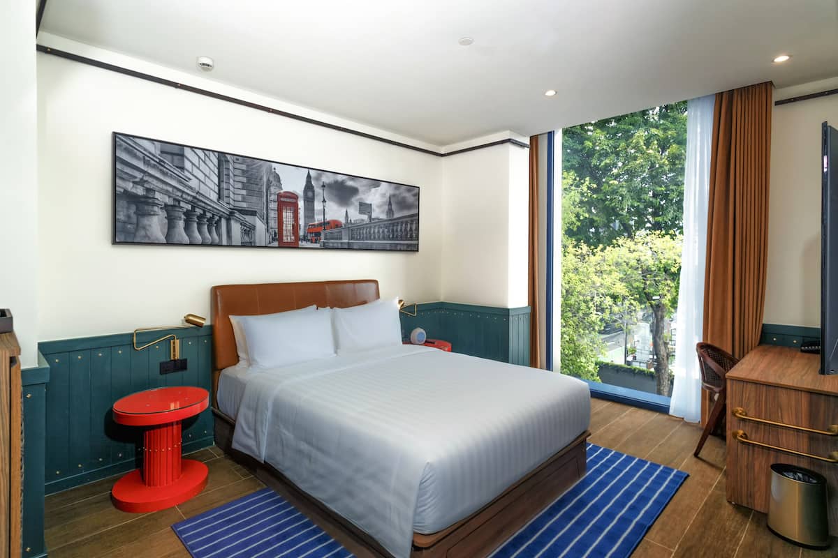 Une chambre d’hôtel avec un grand lit, une table de chevet rouge, un tableau représentant un paysage urbain, une vue sur les arbres par la fenêtre et une commode en bois.