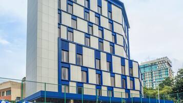 Un edificio moderno con fachada azul y blanca, una cancha de tenis al frente y una valla verde rodeando la cancha.