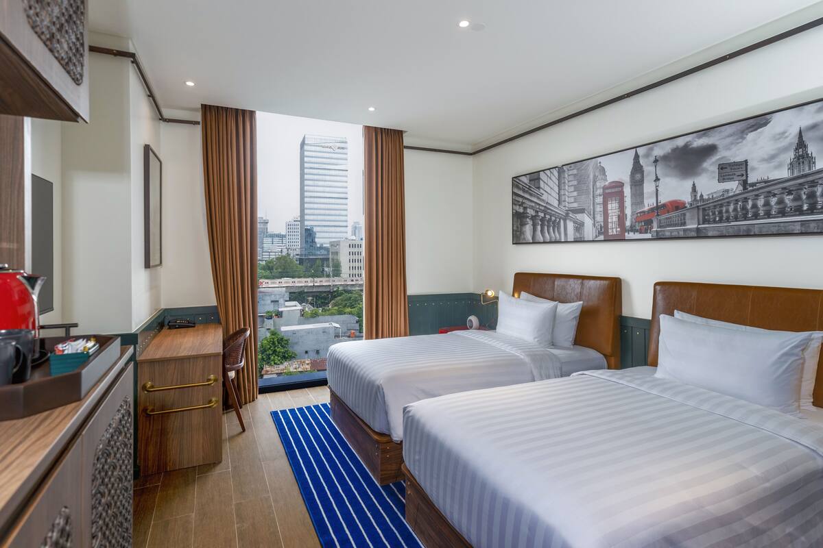 Une chambre d’hôtel avec deux lits, un bureau, une chaise, une fenêtre avec des rideaux et une vue sur la ville.