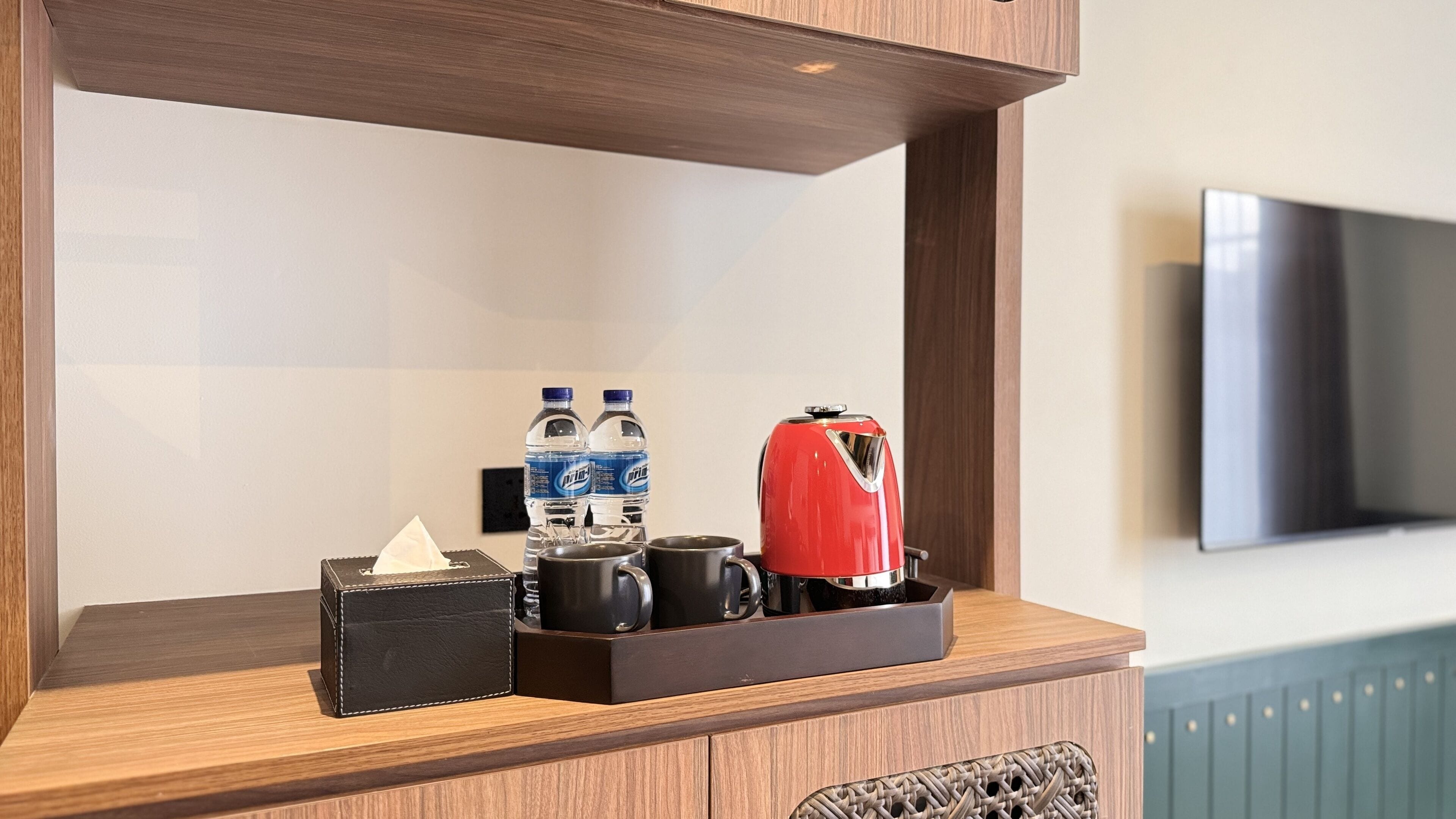 Minibar de hotel con caja de pañuelos, dos botellas de agua, una cafetera roja y una bandeja.