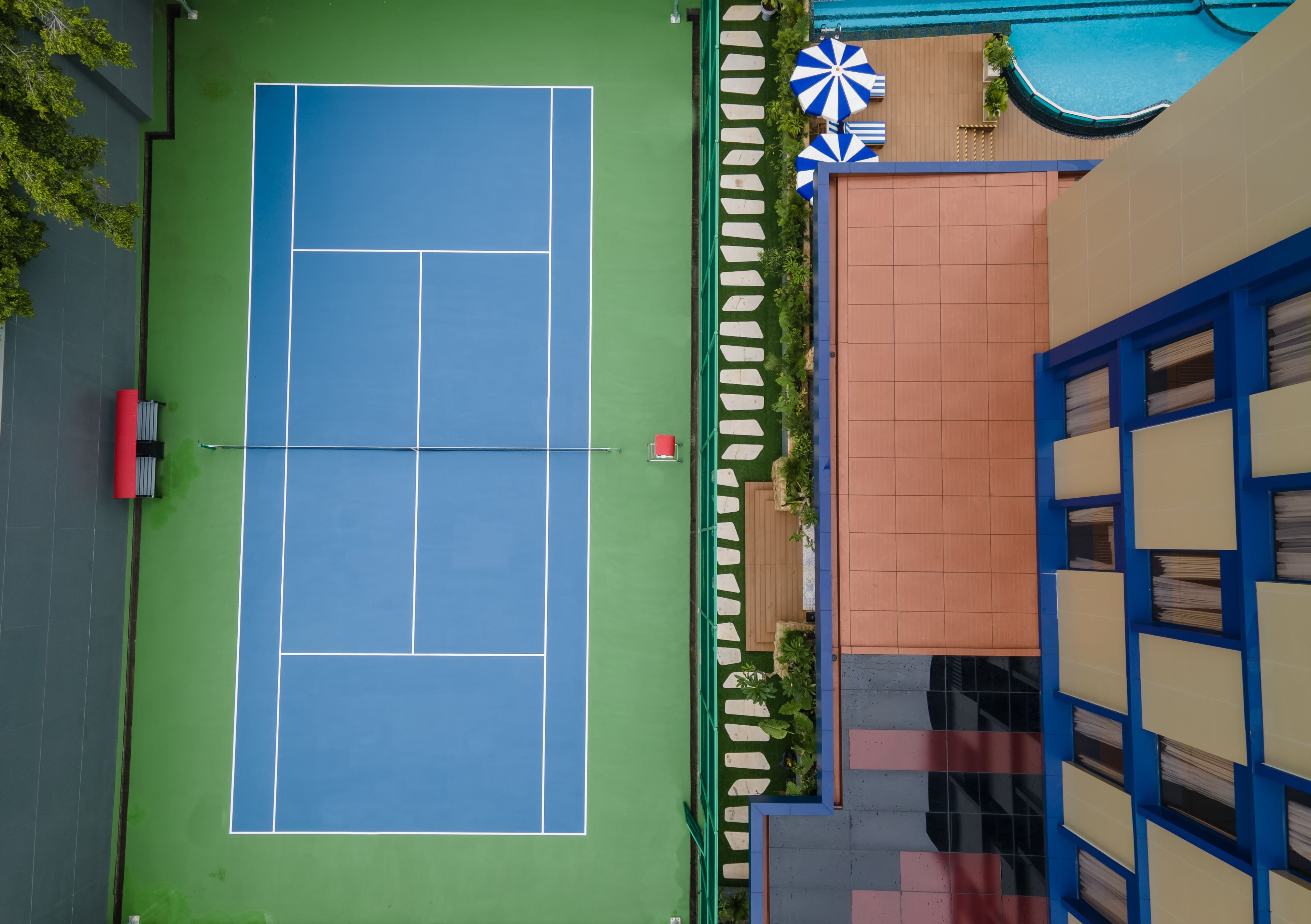 An aerial view of a tennis court with a green surface, adjacent to a building with a blue and beige facade, and a pool area with umbrellas.