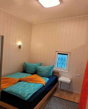 Une petite pièce, de taille modeste, comprenant un lit, une fenêtre, une table de chevet et un luminaire fixé au mur.