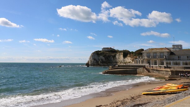 A coastal scene with a sandy beach, rocky cliffs, and a building with a flag. - 1 Star Cottages (Freshwater)