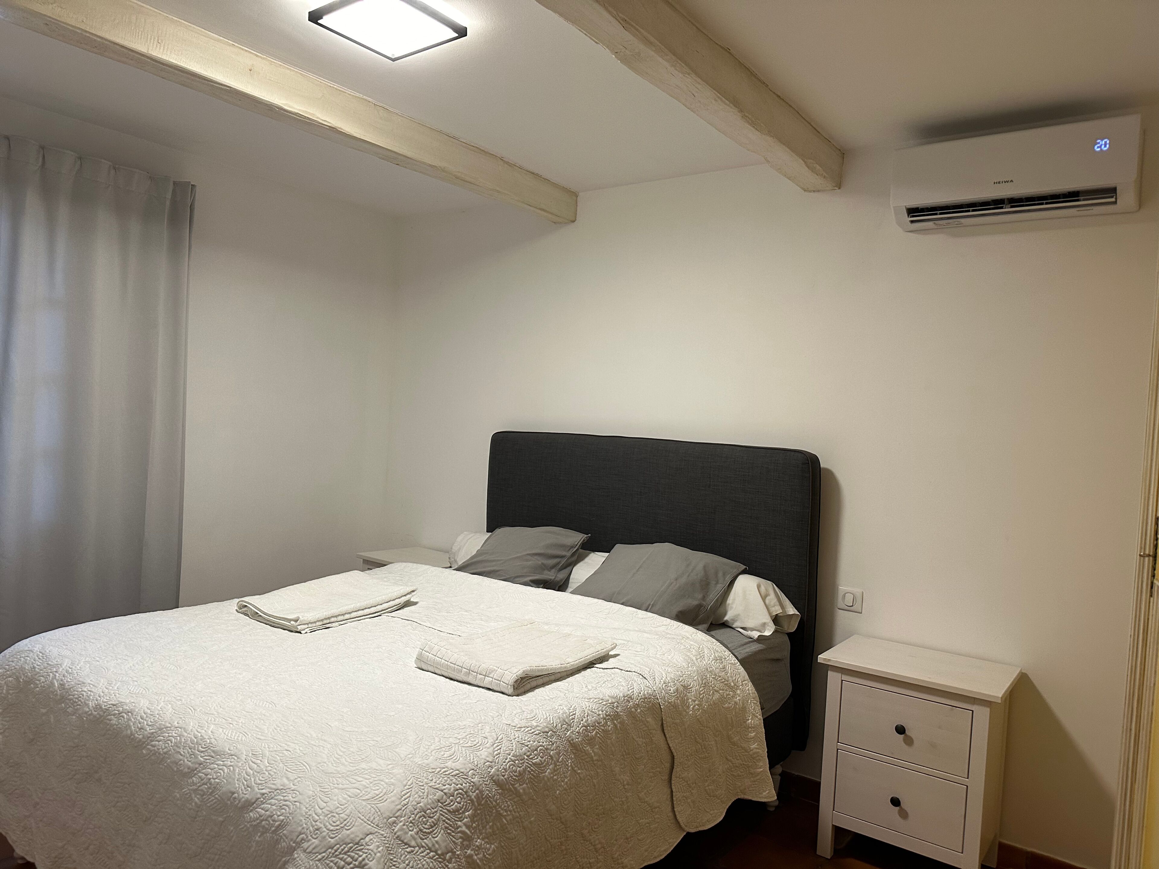 Quarto com cama, mesa de cabeceira, ar-condicionado e janela com cortinas.