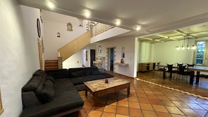 Un vaste espace de vie comprenant un grand canapé d’angle, une table basse en bois et un coin repas avec une longue table et des chaises. Un escalier avec une rampe en bois mène à l’étage supérieur.