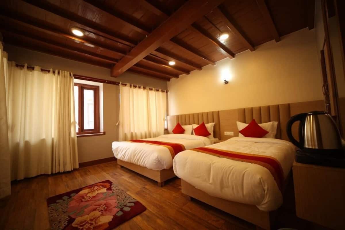 Habitación de hotel con dos camas, vigas de madera en el techo, una ventana con cortinas, un tapete floral y una tetera sobre una mesita.