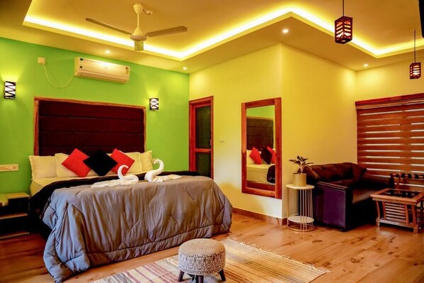 A bedroom with a large bed, a ceiling fan, a small round ottoman, and a wooden side table.