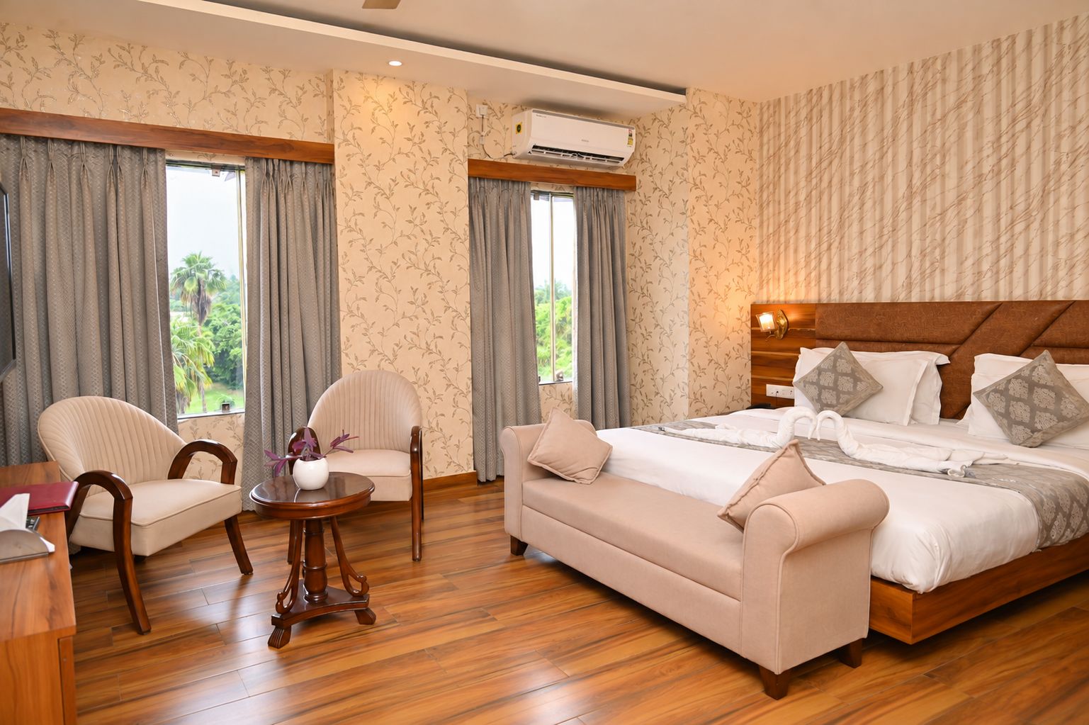 A hotel room with a bed, two armchairs, a small table, and a view of greenery through the windows.
