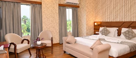 A hotel room with a bed, two armchairs, a small table, and a view of greenery through the windows.