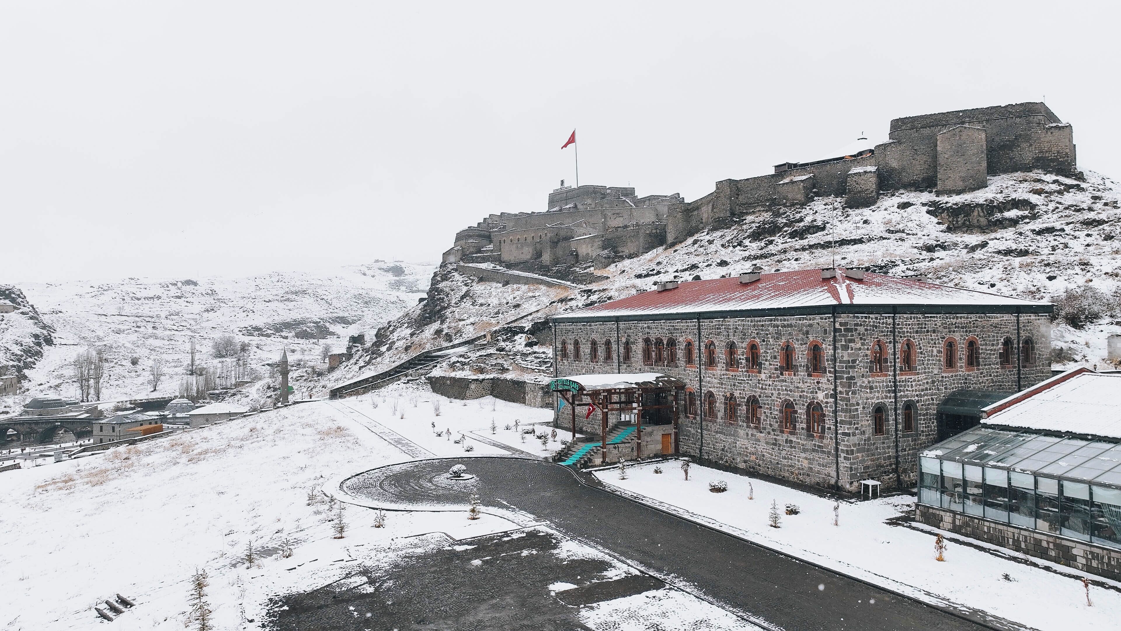 Un edificio histórico de piedra con ventanas arqueadas y techo rojo, rodeado de colinas nevadas y un castillo al fondo.