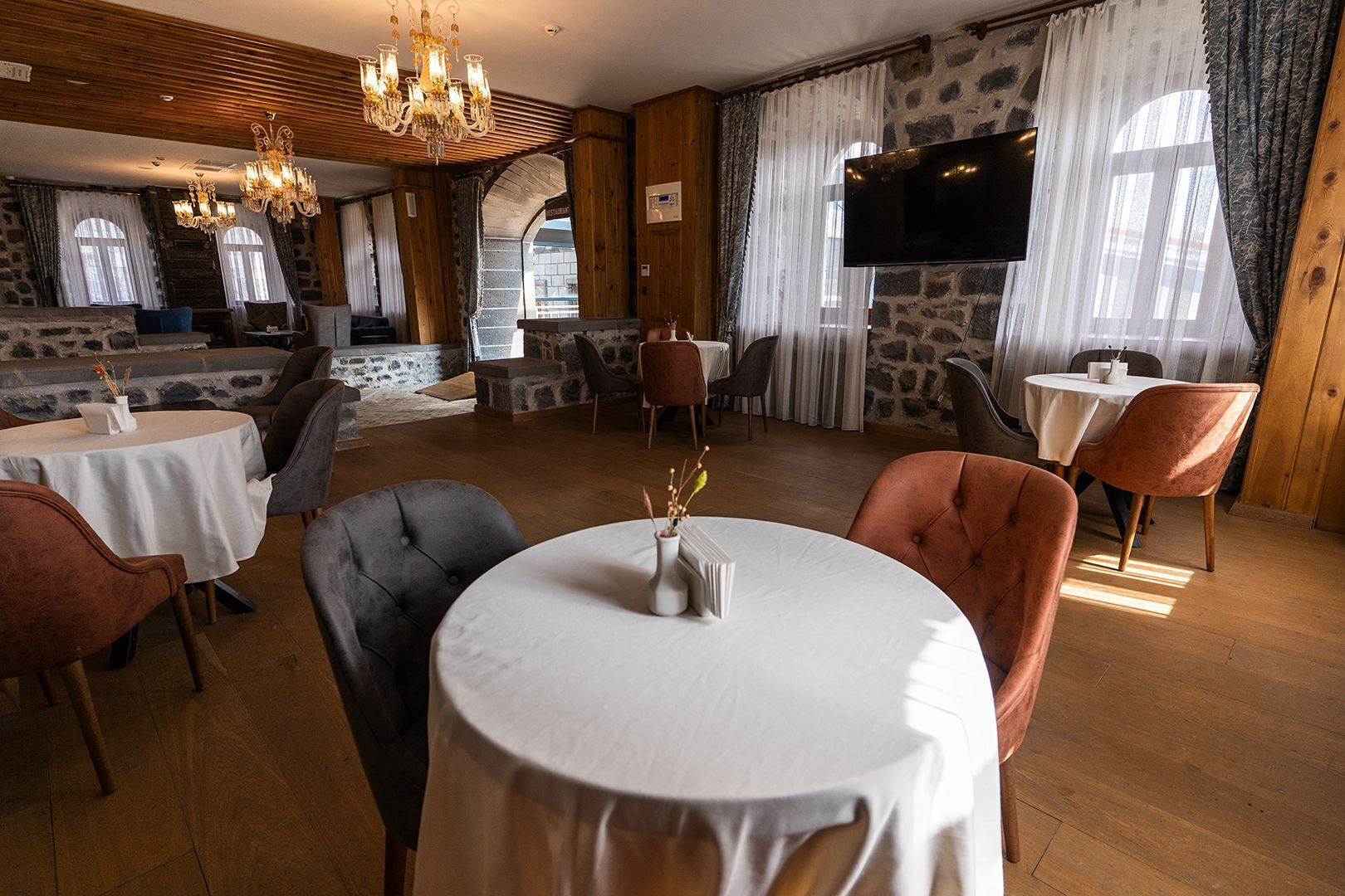 A restaurant with round tables, cushioned chairs, a chandelier, stone walls, and a TV.