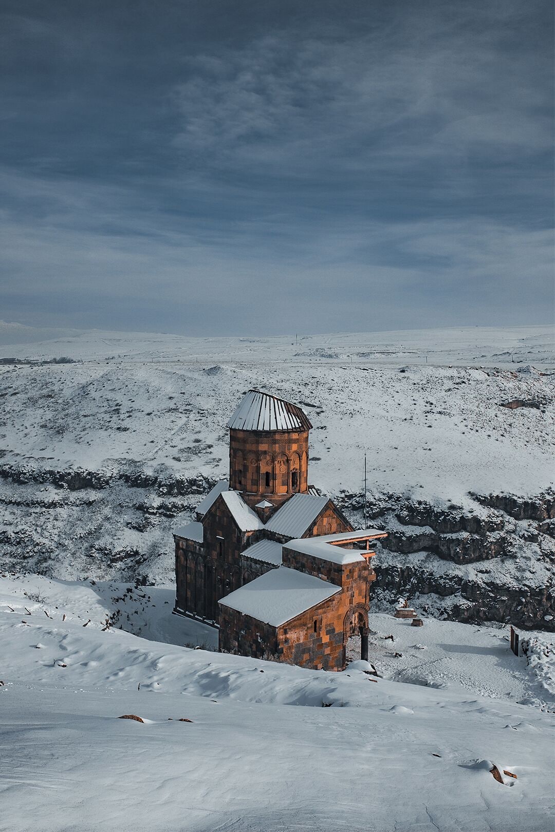 Una iglesia cubierta de nieve con un campanario y un edificio adyacente más pequeño, rodeada por un extenso paisaje nevado.