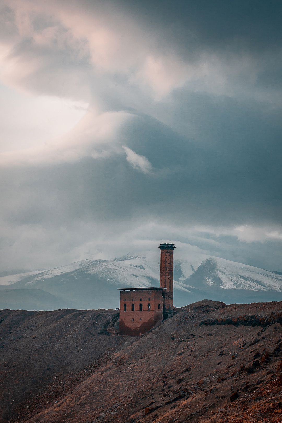 A solitary brick tower on a barren hill with a snow-capped mountain in the background.