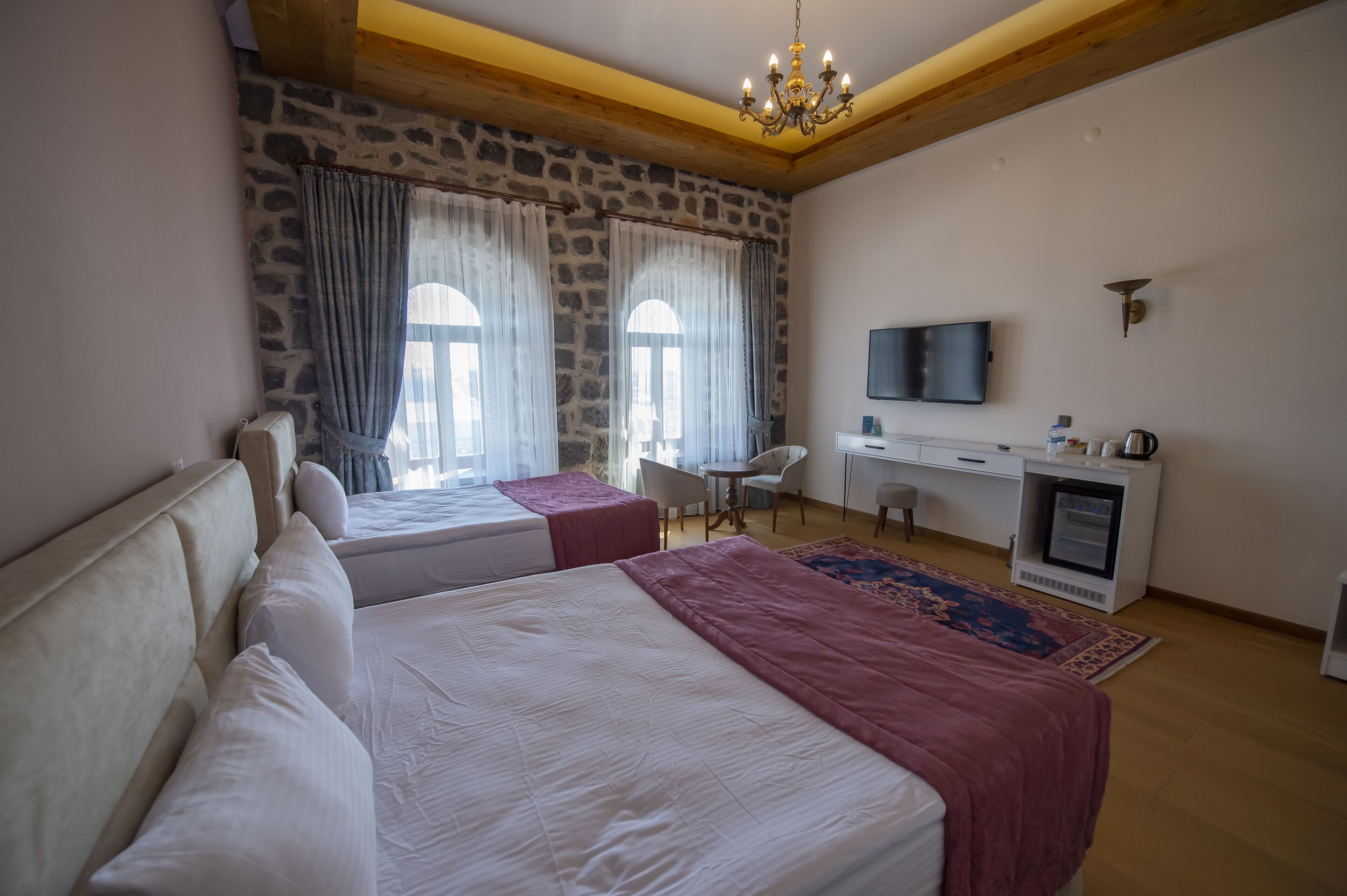 A hotel room with two beds, a desk, a TV, and a stone wall.