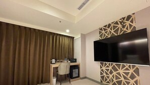 A hotel room with a large flat-screen TV mounted on the wall, a desk with a chair, a small refrigerator, and a curtain-covered window. - SHINSHIN HOTEL (Taipei)