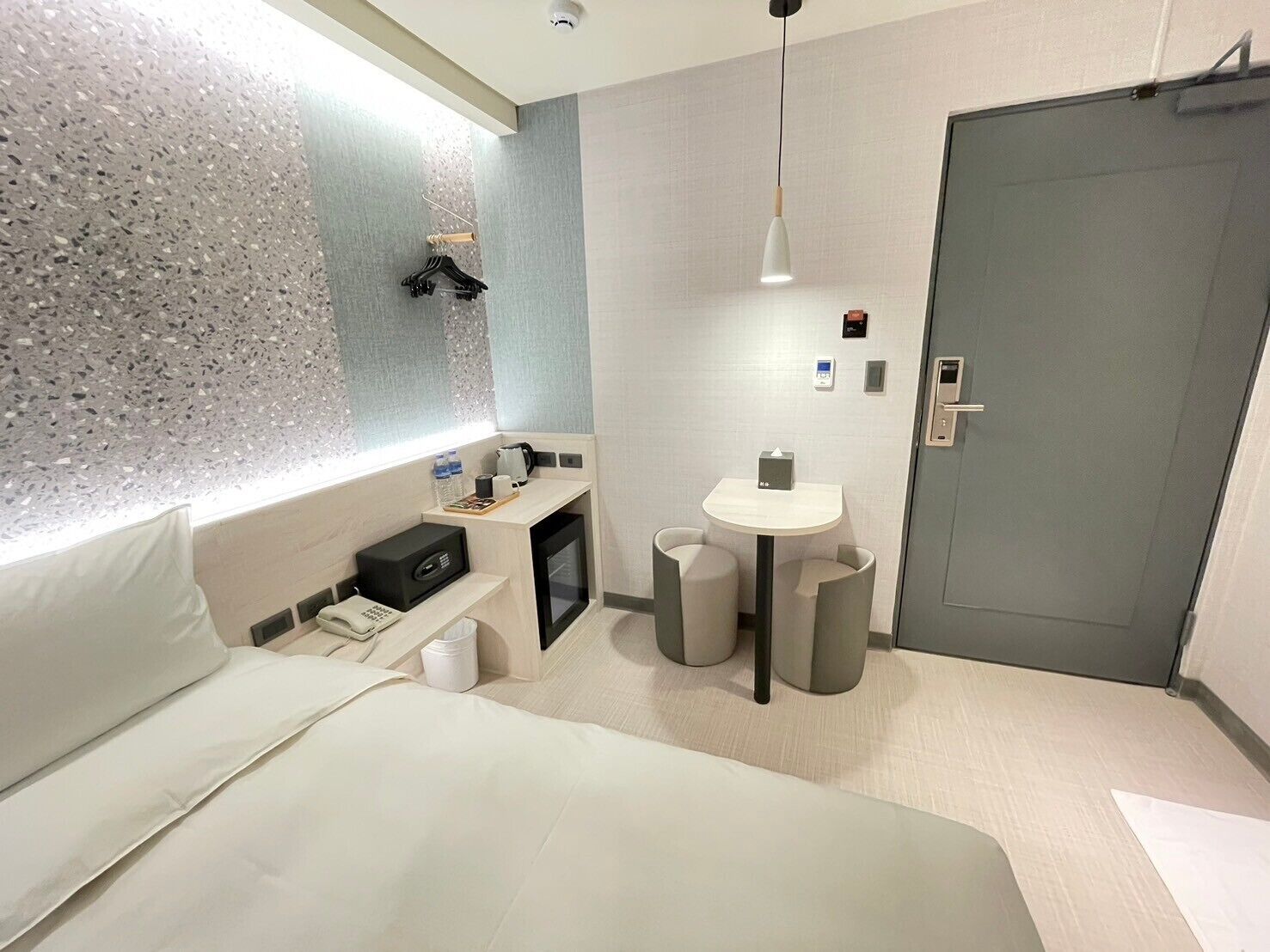A modern hotel room with a bed, bedside table, a small desk with a chair, a round table with two chairs, a wall-mounted heater, and a door.
