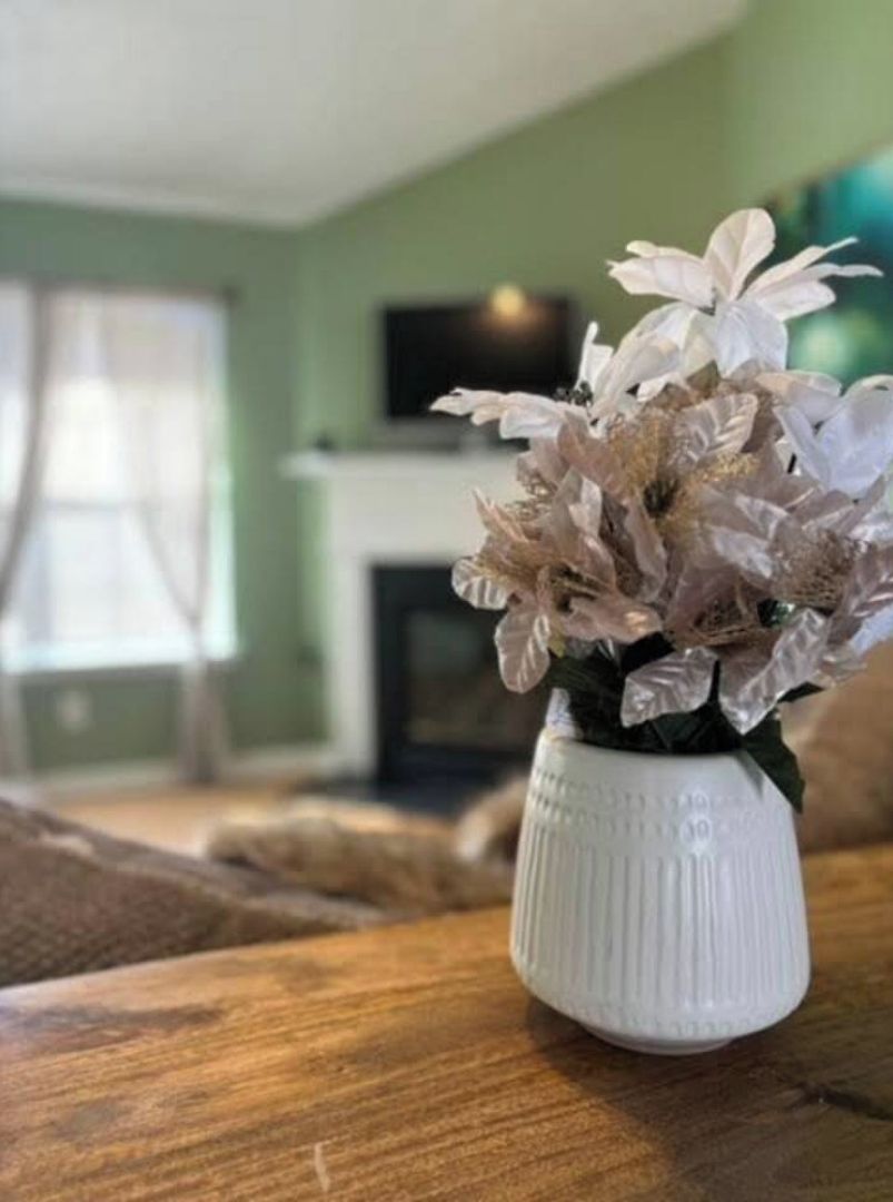 A white vase with a floral arrangement on a wooden table in a room with green walls and a fireplace.