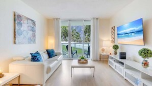 Un salon moderne avec un canapé blanc, une table basse et un téléviseur posé sur un meuble blanc. Une grande fenêtre offre une vue sur des palmiers et un immeuble.