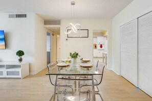 A modern dining area with a glass table and chairs, a decorative wall piece, and a view into the kitchen. - Beachfront Haven: Pools, Direct Ocean Access (Hollywood)