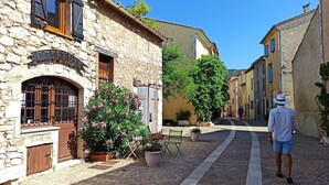 A narrow cobblestone street lined with stone buildings, some with wooden shutters. There are potted plants and a bench along the path.