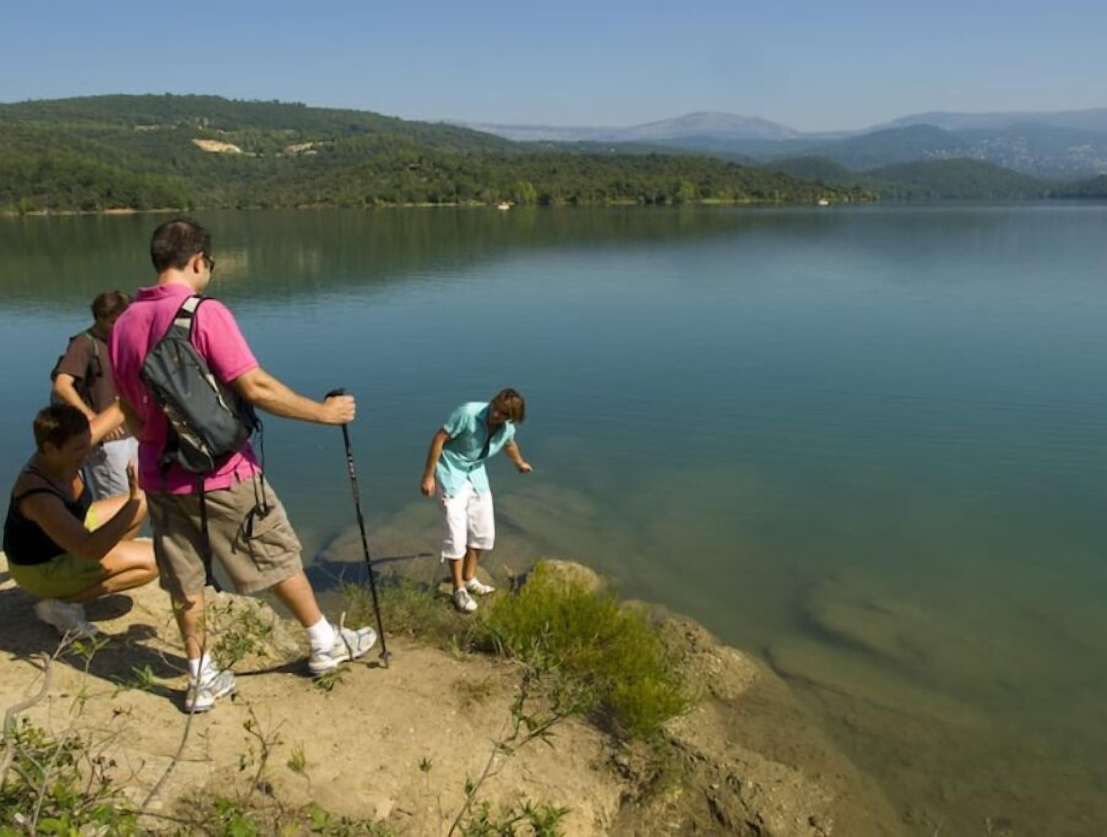 Un groupe de personnes se trouve près d’un lac aux eaux cristallines, entouré de collines et de montagnes en arrière-plan.