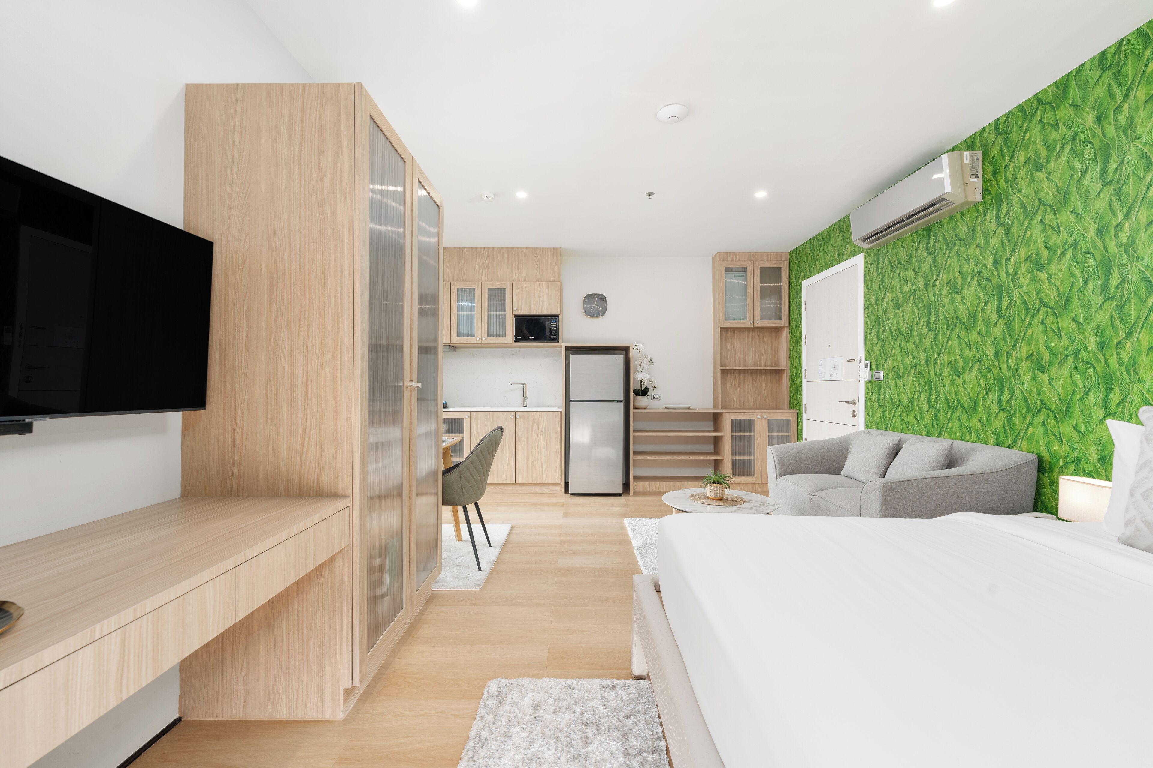 Une chambre moderne avec un lit, une télévision, un canapé gris et un coin cuisine à l’arrière-plan.