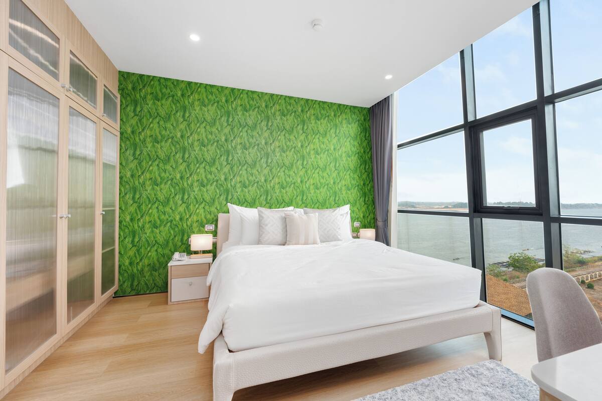 A modern bedroom with a large bed, a nightstand, a chair, and a view of the water through large windows.