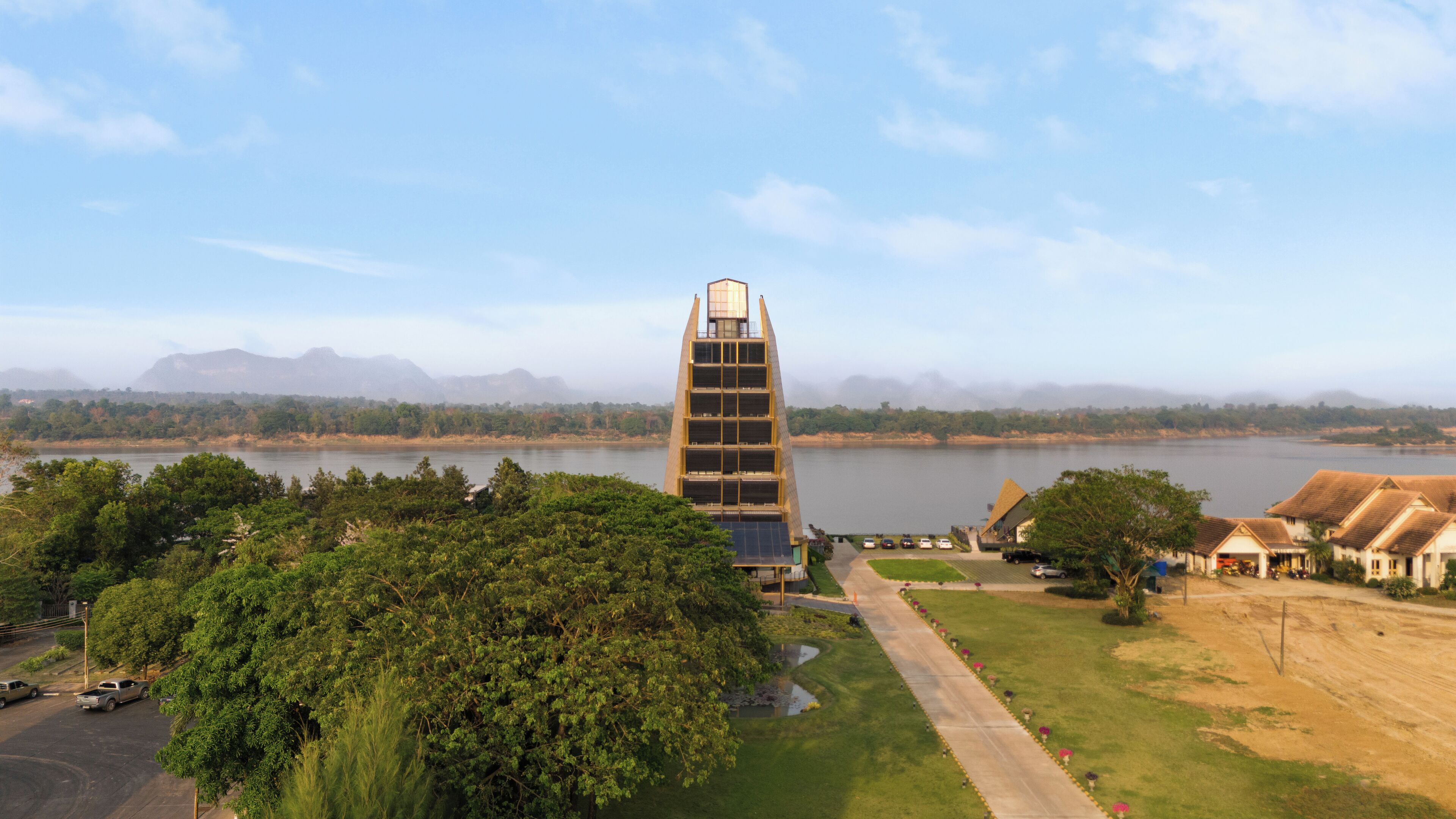 A tall, modern building with a unique design, surrounded by lush greenery and a body of water.