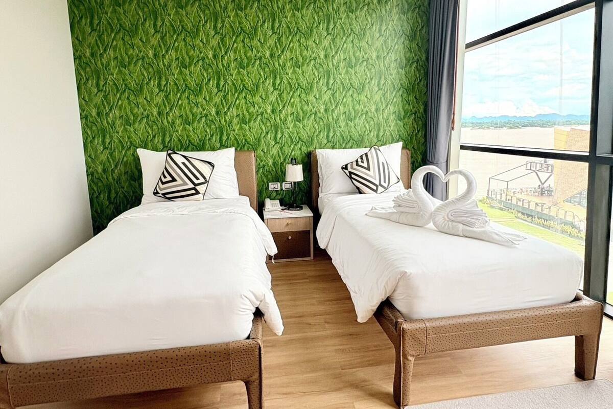 A hotel room with two beds, a green textured wall, a view of a sports field, and a window with curtains.
