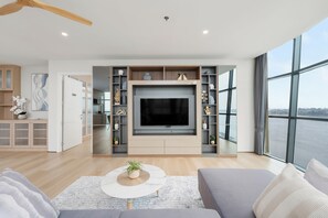 A modern living room with a large flat-screen TV, built-in shelving, and a large window offering a view of the city. - NAKA-RAJ Escape Hotel (Nakhon Phanom)