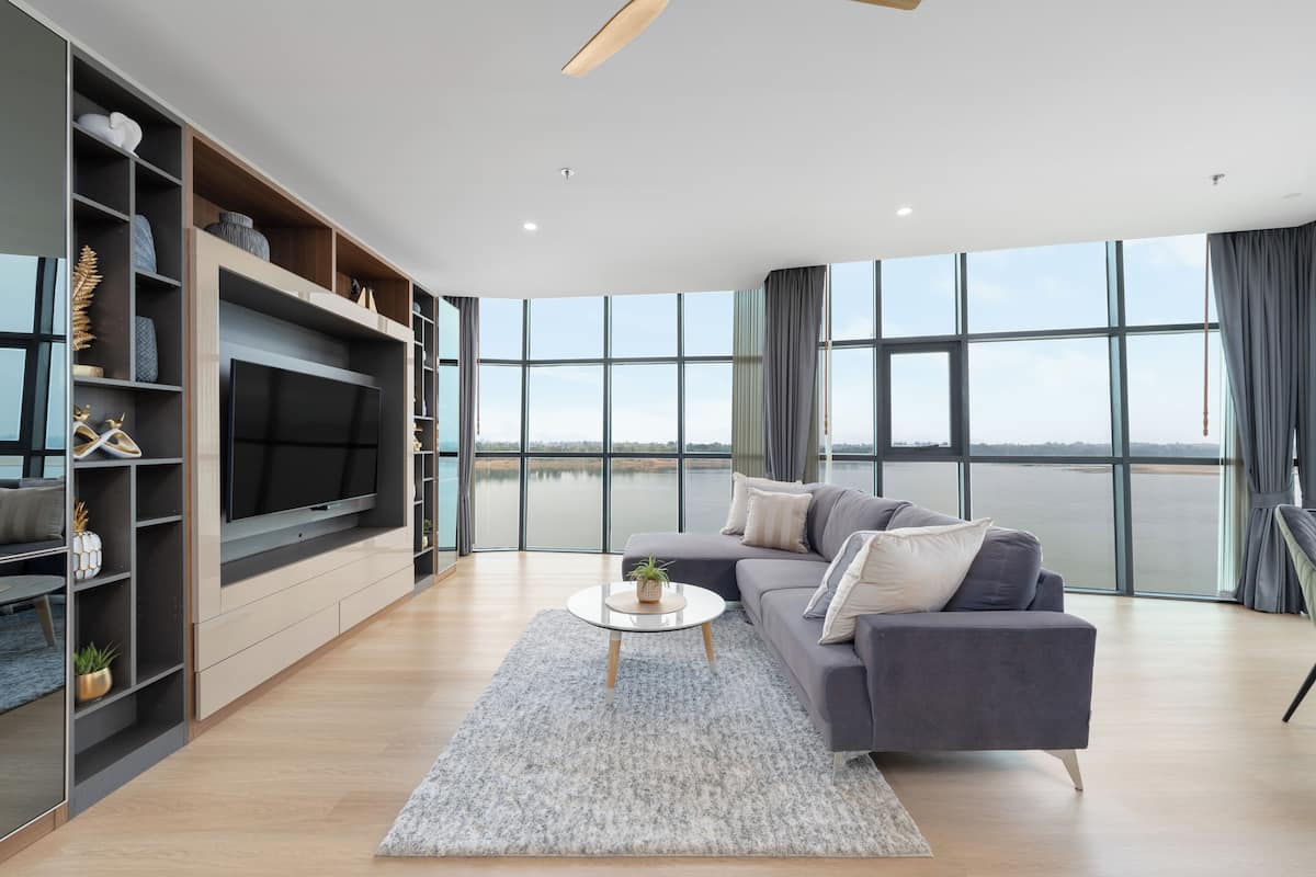 A modern living room with a large flat-screen TV, a grey sofa, and a view of a body of water through large windows.