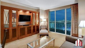 A spacious living room with a large flat-screen TV, a comfortable sofa, a glass coffee table, and a view of the cityscape through large windows.