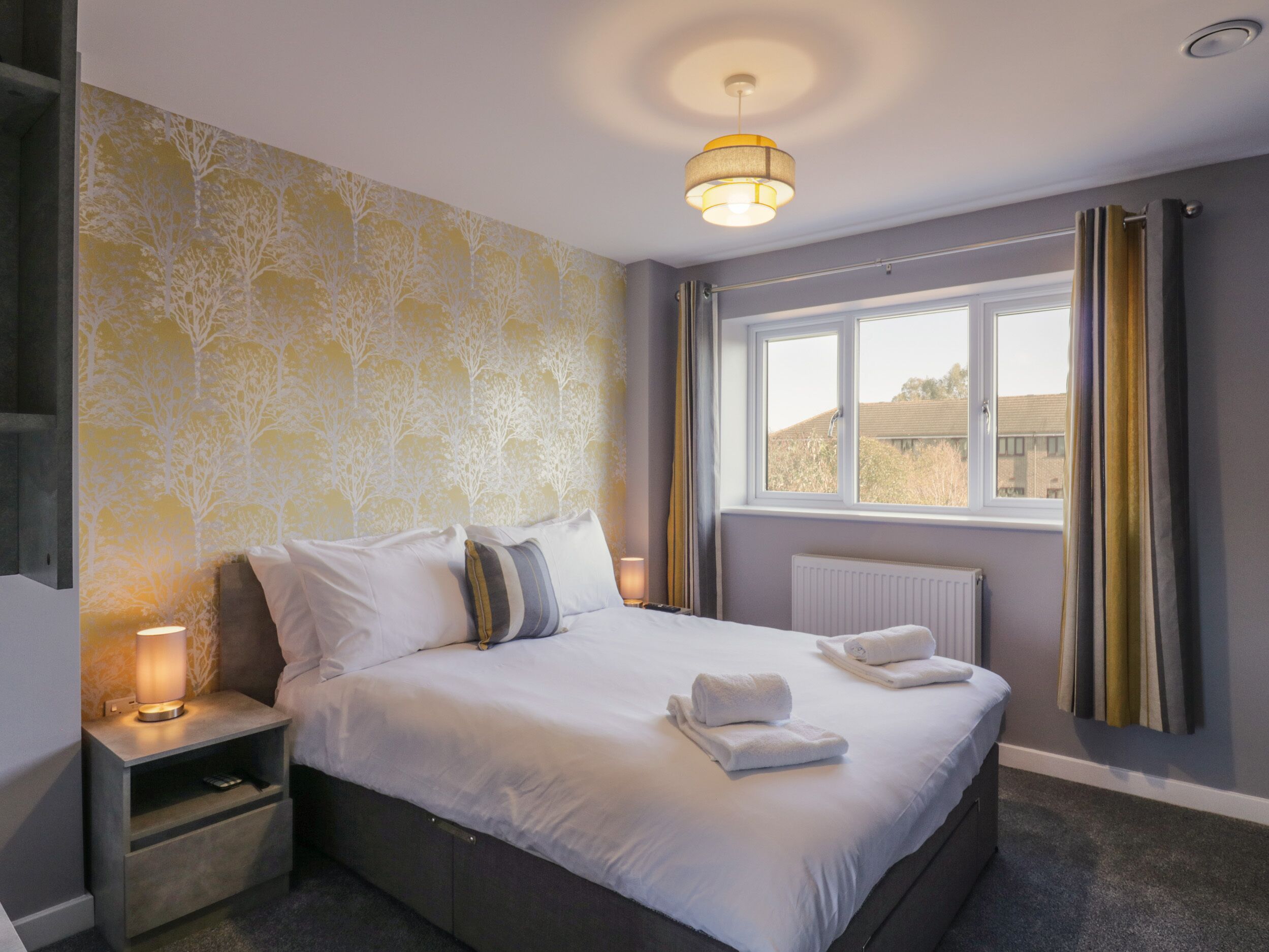 A neatly made bed with white linens and two pillows, a bedside lamp, and a window with curtains.