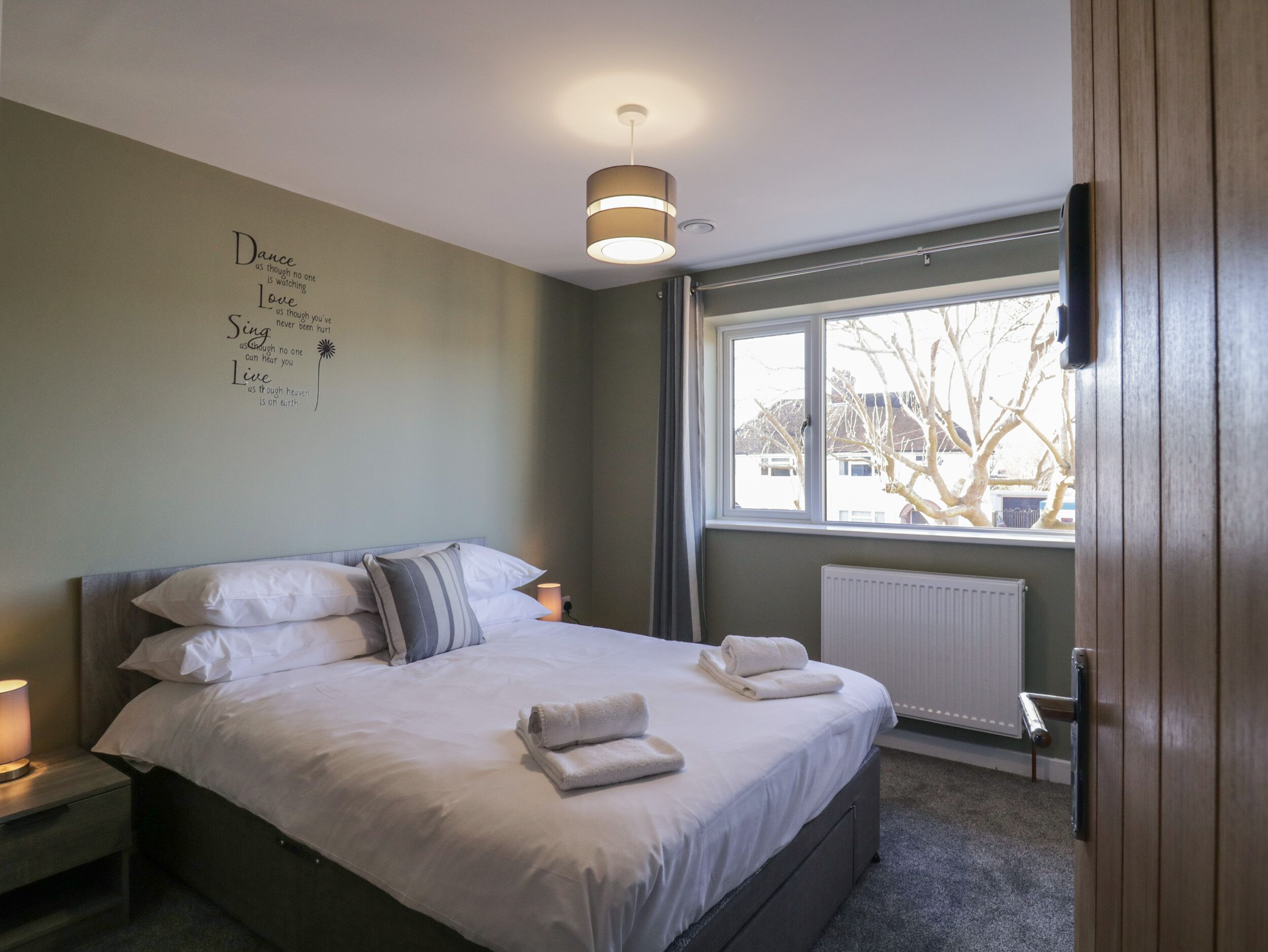 Une chambre avec un lit, une table de chevet, un mur orné de citations inspirantes, une fenêtre donnant sur des arbres et un radiateur.